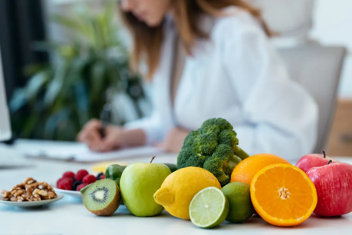 A functional nutritionist writing notes at a bright clinic desk piled with fresh fruits, vegetables, and walnuts — symbolizing food-first, root-cause care